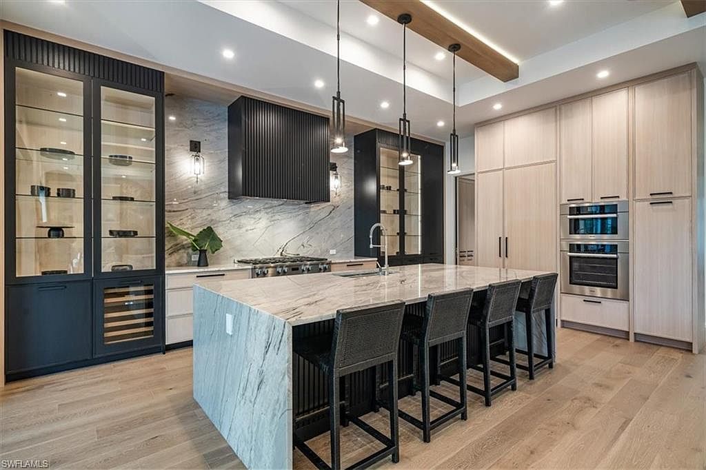 This is a modern kitchen featuring a large island with a marble countertop and seating for five. The kitchen includes custom cabinetry, stainless steel appliances, and pendant lighting. The overall style is contemporary and luxurious, with a focus on clean lines and high-end finishes.