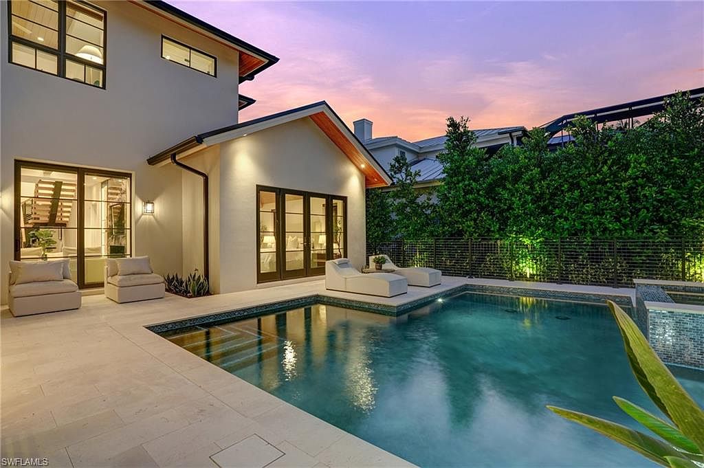 This image showcases a luxurious backyard pool area at dusk, featuring a modern home with large windows and doors that open to the patio. The pool is surrounded by elegant stone tiling and lounge chairs, with lush greenery providing privacy. The scene exudes a sense of relaxation and upscale outdoor living.