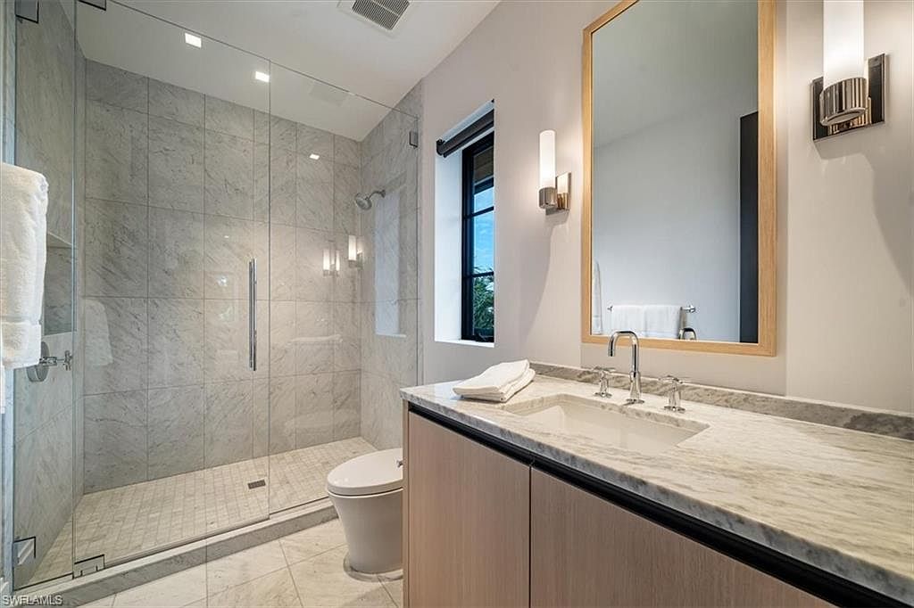 This is a well-lit bathroom featuring a glass-enclosed shower with marble-like tiling, a modern vanity with a light wood finish and marble countertop, and a large mirror. The space is clean and contemporary, with a neutral color palette that creates a serene and inviting atmosphere. The perspective is from the doorway, showcasing the entire bathroom.