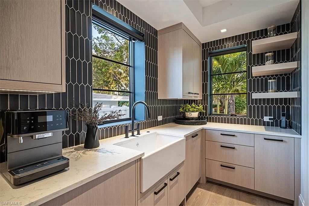This is a well-lit kitchen featuring light wood cabinetry with black hardware, a white farmhouse sink, and black framed windows that offer a view of the outdoors. The backsplash is a unique dark tile pattern, and the countertops are a light, neutral color. The overall impression is modern and stylish.