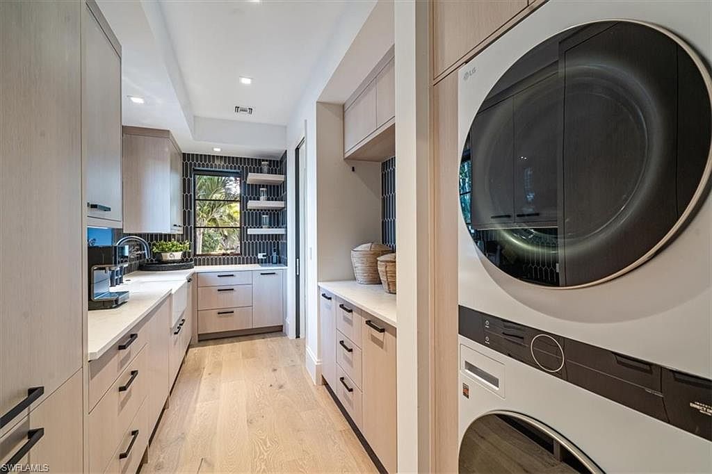 This is a modern laundry room featuring light wood cabinetry with sleek black hardware, light wood flooring, and a stacked washer and dryer unit. A window provides natural light and a view to the outside, while decorative baskets add a touch of warmth to the space. The overall impression is clean, organized, and stylish.