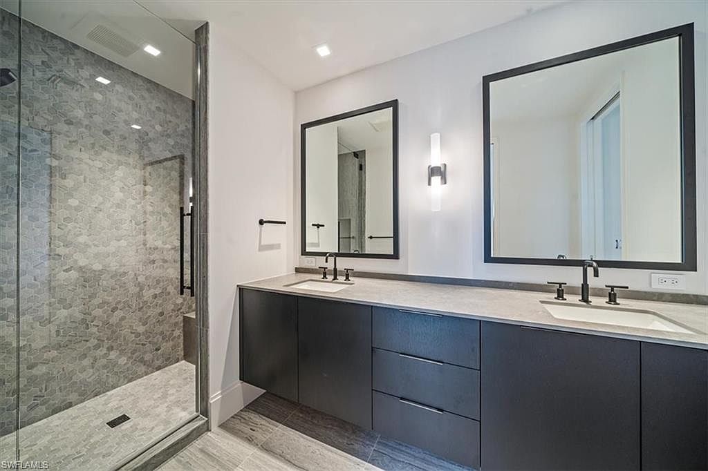 This is a modern primary bathroom featuring a double vanity with dark cabinetry and light countertops. Two square mirrors with black frames hang above the sinks, illuminated by a sleek sconce. A glass-enclosed shower with gray hexagonal tiles adds a luxurious touch, and the overall design is clean and contemporary.
