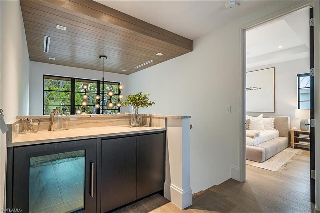 This interior shot showcases a stylish hallway leading into a bedroom. The hallway features a built-in bar area with dark cabinetry and a light countertop, complemented by a modern light fixture. The bedroom is visible through an open doorway, revealing a neutral color palette and a minimalist design.