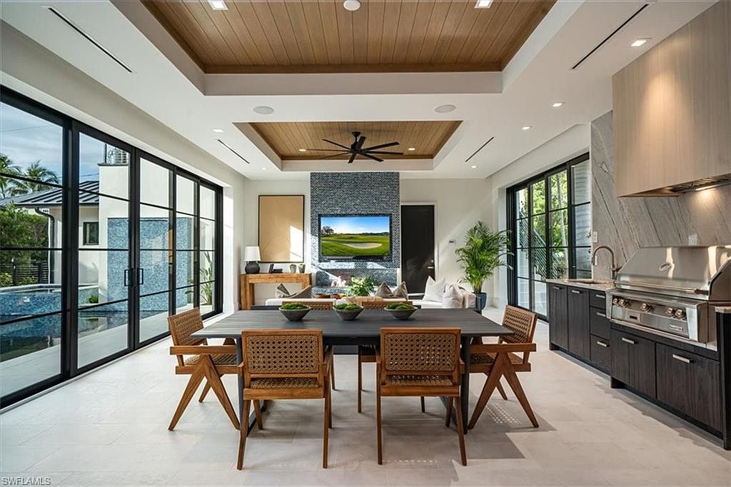 This interior shot showcases a modern dining room with a dark wood table surrounded by woven chairs. Large sliding glass doors provide ample natural light and views of the exterior, while a stone accent wall with a television adds a contemporary touch. The space seamlessly blends dining and living areas, creating an open and inviting atmosphere.