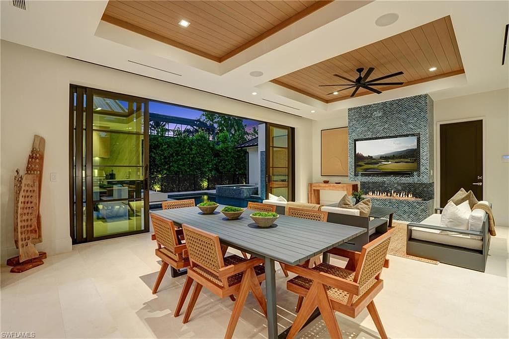 This interior shot showcases a modern dining room with a large table set for six, featuring a sleek gray tabletop and woven chairs. The room opens to an outdoor patio with a pool through large sliding glass doors, enhancing the indoor-outdoor living experience. A unique fireplace with a blue tile surround and a mounted television adds a focal point to the space.