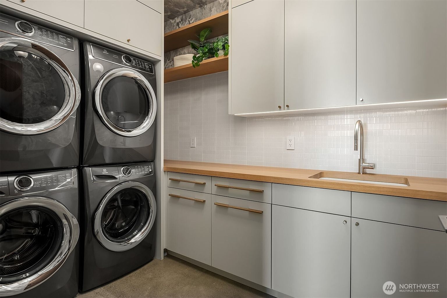 This modern laundry room features a stacked washer and dryer set next to a sleek, light-toned cabinetry unit with a light wood countertop and an integrated sink. The space is accented by a white tiled backsplash, open shelving with greenery, and minimalist hardware, creating a clean and functional aesthetic. The perspective is a straight-on shot that highlights the efficient use of space and contemporary design elements.