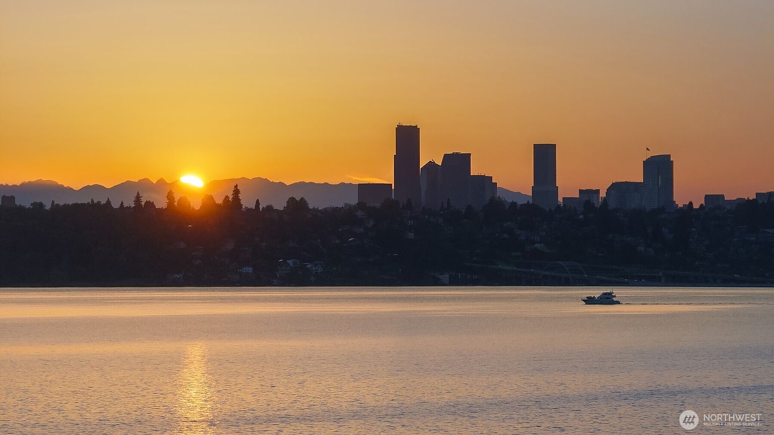 This scenic landscape shot captures a panoramic view of a city skyline silhouetted against a vibrant, golden sunset. In the foreground, a calm body of water reflects the warm light, with a small boat cruising across the surface. The composition emphasizes the expansive horizon and the peaceful atmosphere of the waterfront setting.
