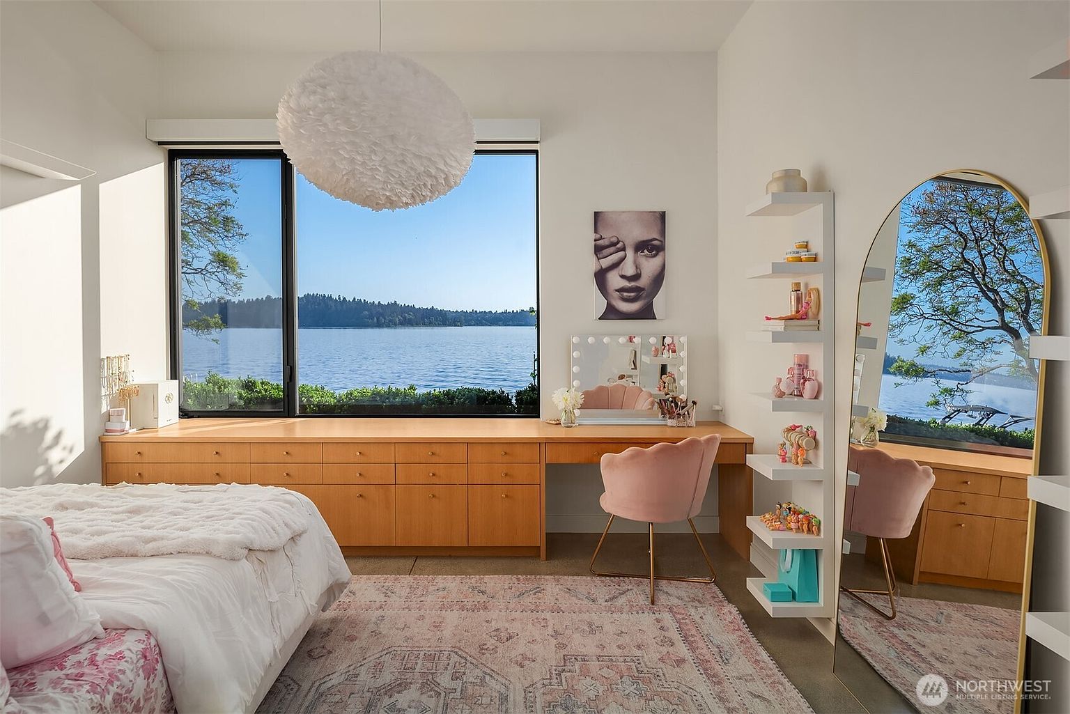 This bright and airy bedroom features a large window overlooking a serene lake, providing a stunning natural backdrop. The room is furnished with a long, light wood desk unit that doubles as a vanity, complemented by a soft pink chair and a decorative wall-mounted shelving unit. A plush bed with a white textured comforter and a patterned area rug complete the cozy, feminine aesthetic of the space.