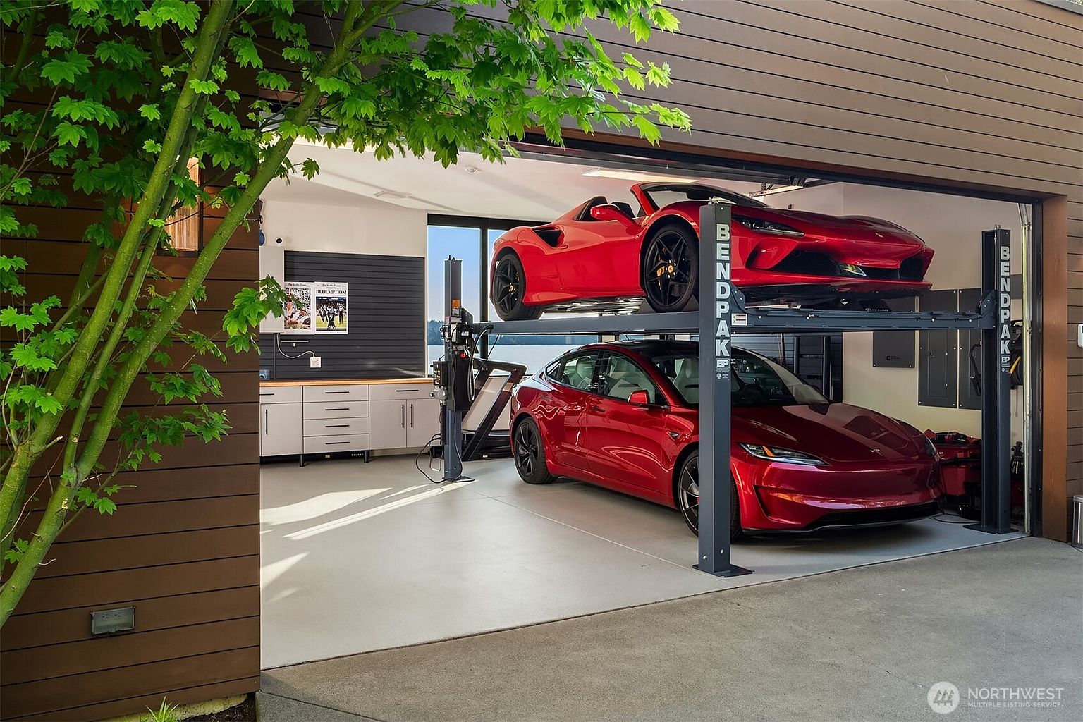 This modern garage features a high-end automotive lift system, showcasing a red sports car elevated above a red electric sedan. The space is finished with clean, light-colored flooring, white cabinetry, and a dark-toned slat wall, creating a sleek and organized environment. A large opening provides a view of the outdoors, framed by contemporary wood siding and a lush green tree in the foreground.