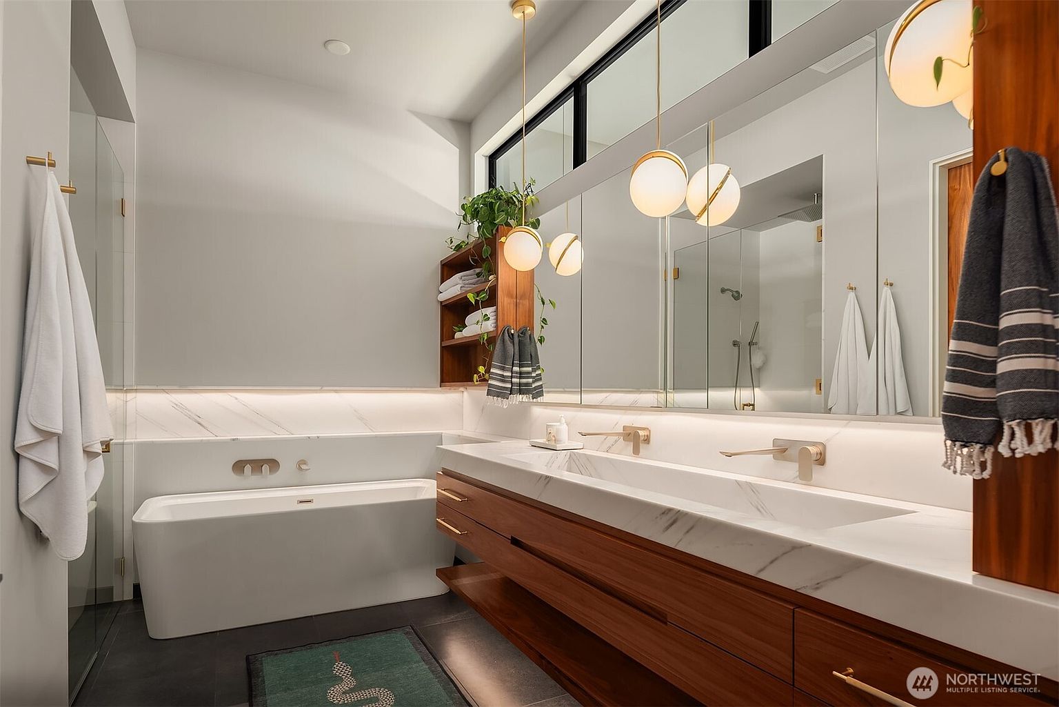 This modern primary bathroom features a sleek, double-sink vanity with a wood-toned base and a stone countertop, complemented by a deep soaking tub set against a wall with integrated lighting. A large mirror spans the vanity area, reflecting the minimalist aesthetic and the warm glow of pendant lights hanging above. The space is accented by a green rug with a snake motif and open shelving filled with towels and greenery, creating a sophisticated and serene atmosphere.