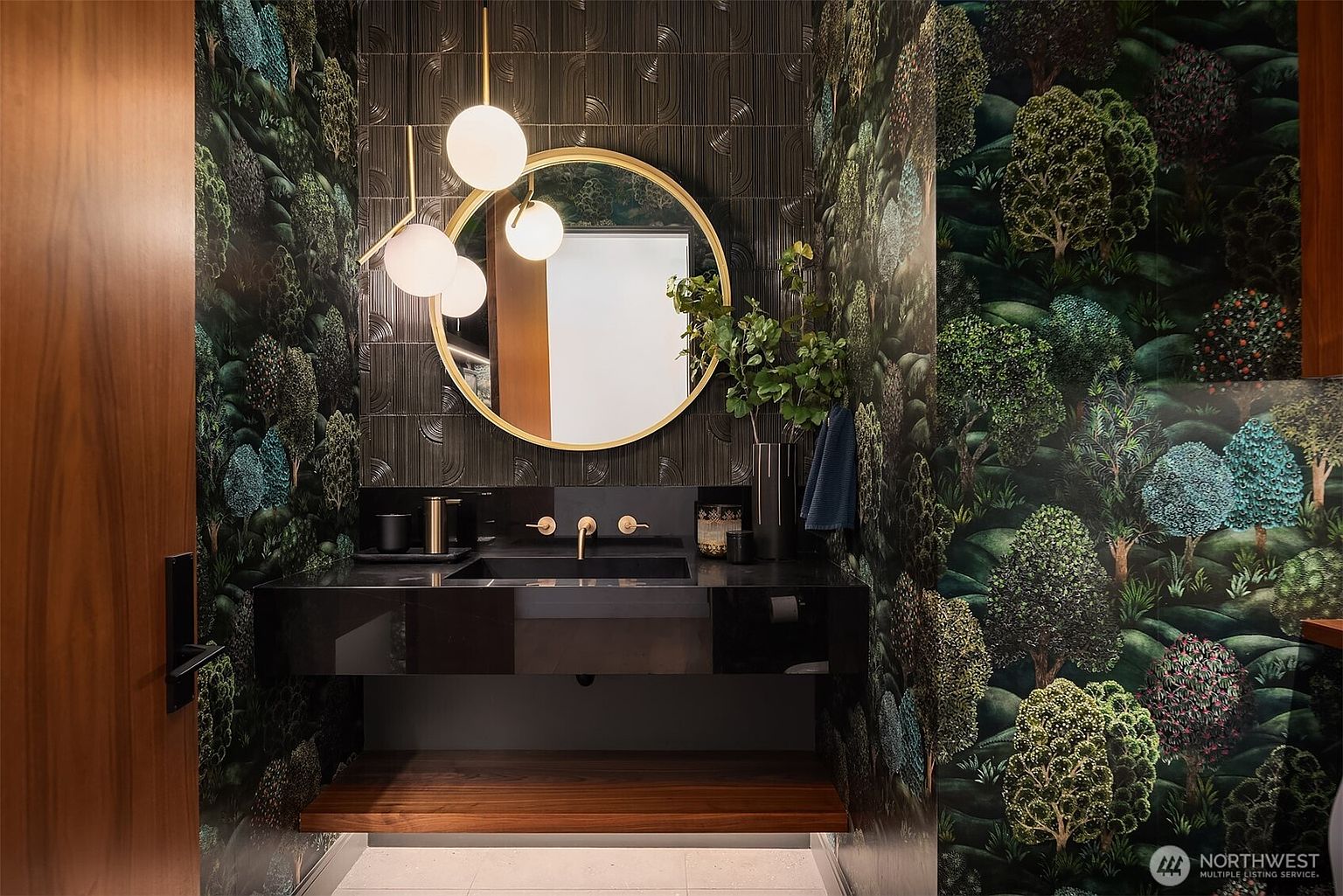 This sophisticated powder room features bold, forest-themed wallpaper and a dark, textured accent wall behind a floating vanity. A circular gold-rimmed mirror is illuminated by a modern, multi-globe pendant light, while a natural wood shelf provides open storage below the sleek black sink. The space offers a moody, high-end aesthetic with a focus on dramatic textures and warm metallic accents.