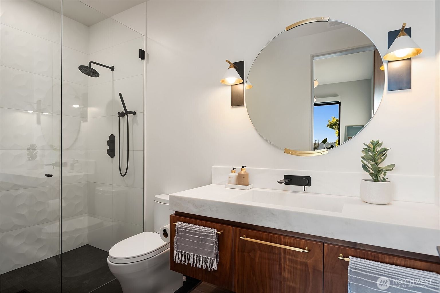 This modern bathroom features a floating vanity with a wood-toned finish, topped with a white stone counter and a wall-mounted faucet. A large, circular mirror with gold-toned accents hangs above the vanity, flanked by two contemporary wall sconces. To the left, a glass-enclosed walk-in shower showcases textured white wall tiles and matte black fixtures, creating a clean and sophisticated aesthetic.
