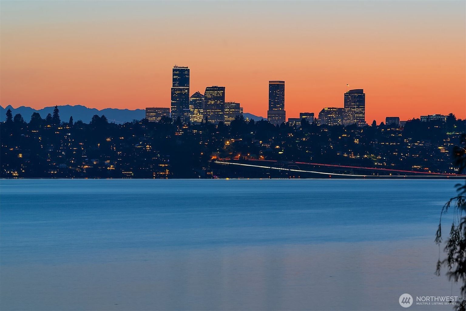 This cinematic wide-angle shot captures a stunning twilight cityscape of Bellevue, Washington, silhouetted against a vibrant orange and purple sunset. The foreground features the calm, reflective waters of Lake Washington, while the mid-ground shows the illuminated skyline and the bridge connecting the shores. The perspective is elevated and distant, emphasizing the serene atmosphere and the prime location of the property's surroundings.