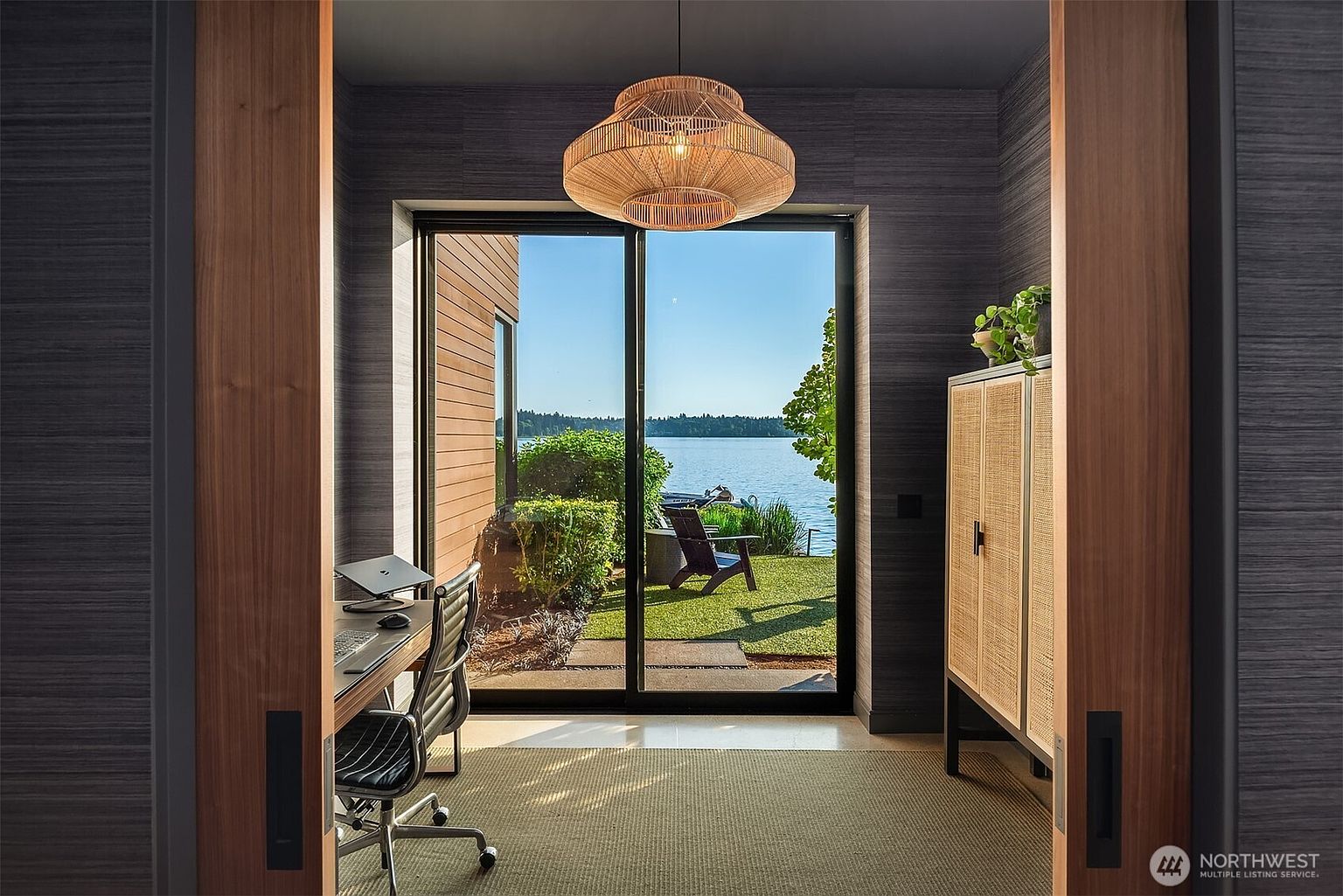 This sophisticated home office features a minimalist desk setup and a tall, textured cabinet with woven door panels, framed by warm wood-paneled walls. A large, woven pendant light hangs centrally, drawing the eye toward the expansive sliding glass doors that provide a seamless transition to the outdoor patio and waterfront view. The room is bathed in natural light, creating a serene and productive atmosphere with a direct connection to the tranquil lake landscape.