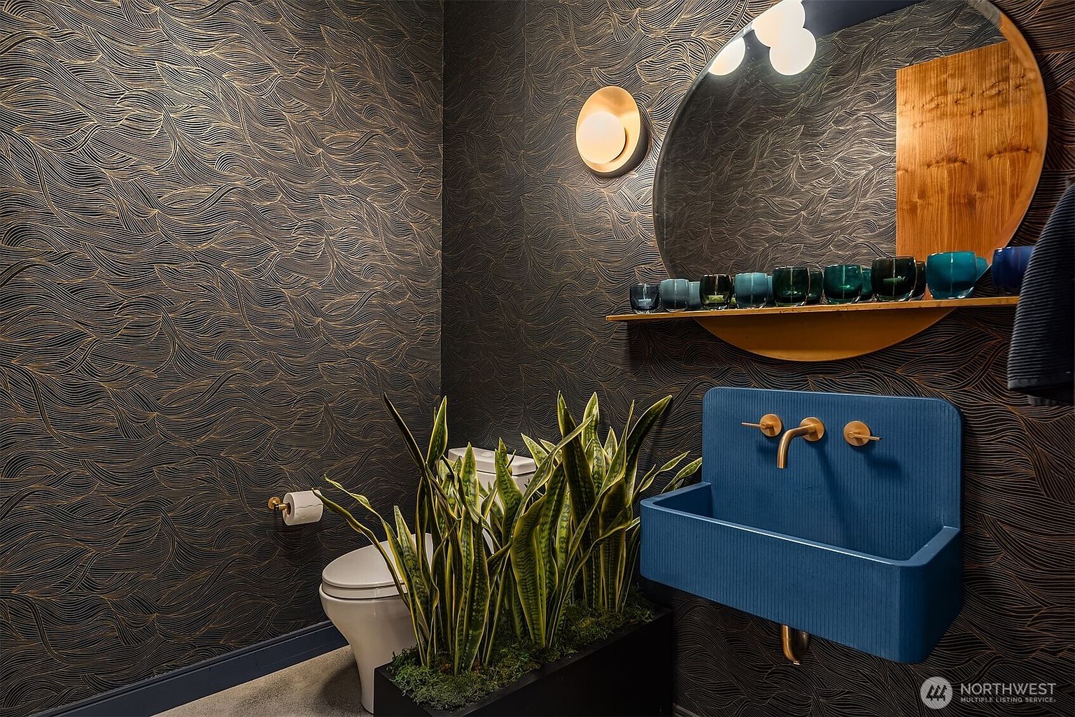 This unique powder room features bold, dark patterned wallpaper and a striking blue wall-mounted sink with gold-toned fixtures. A large circular mirror hangs above a wooden shelf lined with colorful glass tumblers, while a large planter filled with snake plants adds a vibrant touch of greenery. The space is illuminated by a warm, circular wall sconce, creating a moody and sophisticated atmosphere.
