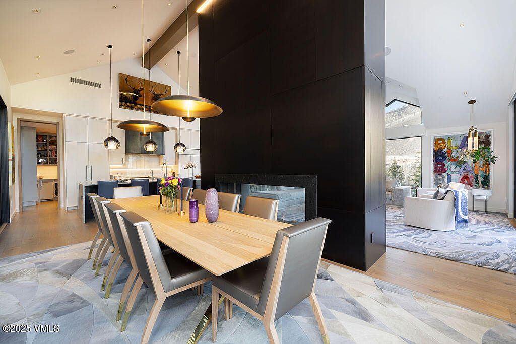 This open-concept dining area features a long light wood table surrounded by modern chairs with grey upholstery. A prominent, dark-paneled central structure separates the dining space from the adjacent living area, while a series of contemporary pendant lights hangs above the table. The room is finished with light wood flooring and a large, patterned area rug, creating a sophisticated and airy atmosphere.