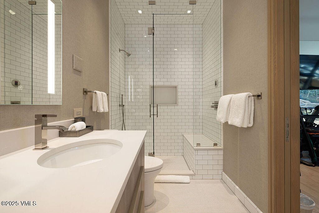 This modern bathroom features a sleek vanity with a white countertop and a rectangular undermount sink, complemented by a tall, integrated light fixture on the mirror. The walk-in shower is enclosed by glass panels and finished with white subway tile, including a built-in niche and a small bench seat. The space is tied together with light-toned wall coverings and small-scale mosaic floor tiles, creating a clean and contemporary aesthetic.