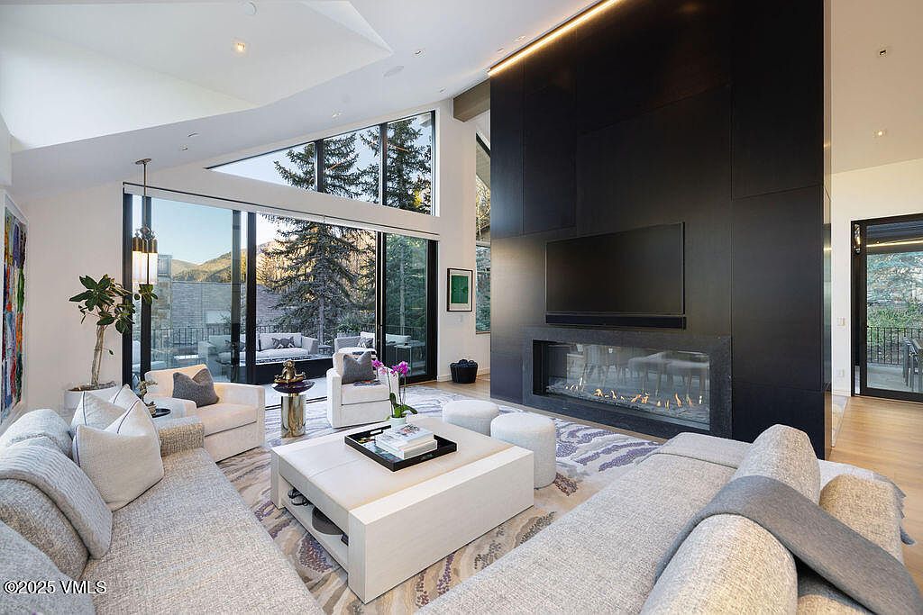 This spacious living room features a contemporary design with a large, dark, full-height fireplace surround that serves as a dramatic focal point. The room is anchored by a light-colored sectional sofa and a central coffee table, all set upon a patterned area rug. Large windows and glass doors provide an expansive view of the outdoor landscape, filling the room with natural light and creating a seamless connection to the exterior.