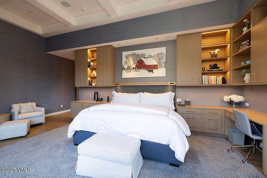 This primary bedroom features a sophisticated design with a blue-toned textured wall and a central bed flanked by custom light wood cabinetry and integrated shelving. A desk area is seamlessly incorporated into the cabinetry on the right, while a cozy armchair and ottoman sit in the corner. The room is anchored by a large, plush area rug and illuminated by soft, warm accent lighting.