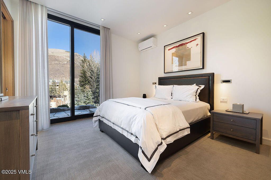 This modern bedroom features a neutral color palette with a dark-framed bed and matching nightstand. A large glass sliding door provides access to the outdoors and offers a scenic view of the surrounding trees and mountains. The room is accented by recessed lighting, a wall-mounted air conditioning unit, and a piece of abstract art above the headboard.
