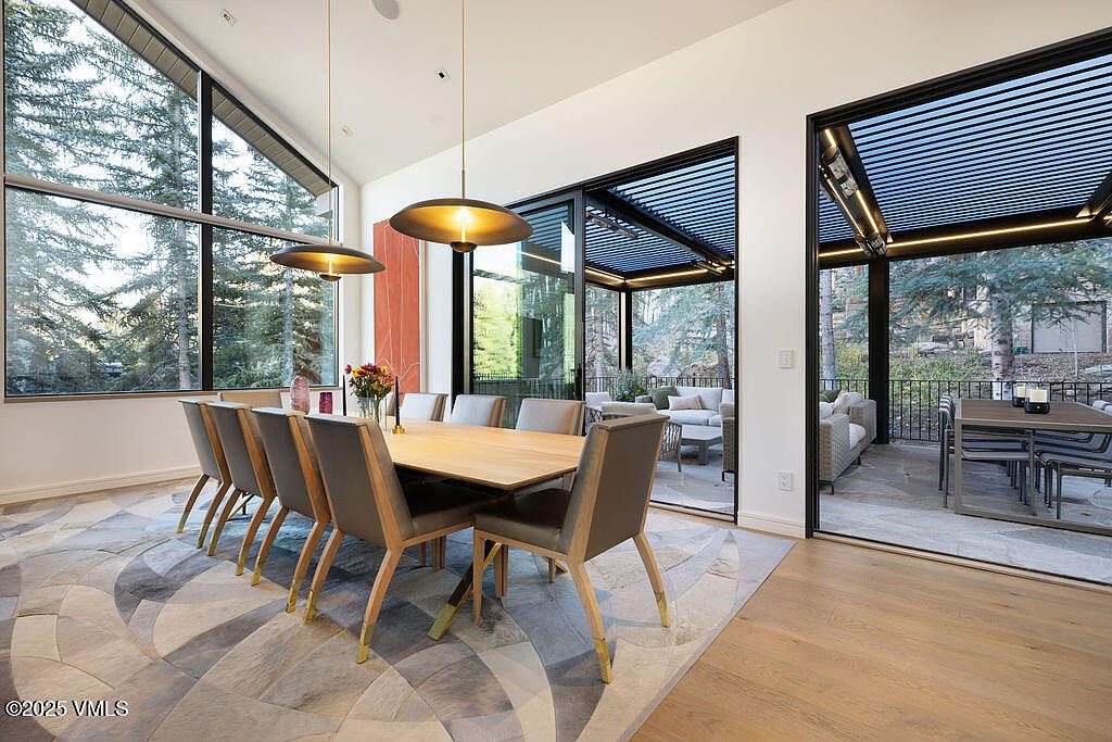 This elegant dining area features a long light wood table surrounded by eight modern, upholstered chairs set atop a large, patterned area rug. The space is illuminated by two sleek, low-hanging pendant lights and benefits from expansive windows that provide a clear view of the surrounding forest. Large sliding glass doors open directly onto a covered outdoor patio, creating a seamless transition between the indoor dining space and the exterior living area.