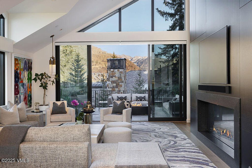 This sophisticated living room features a neutral color palette with a large, textured sectional sofa and two armchairs arranged around a central coffee table. A tall, dark-toned fireplace surround anchors the right side of the room, while a large, colorful abstract painting adds a vibrant focal point on the left. The space opens through expansive glass sliding doors to a mountain-view patio, creating a seamless transition between the refined interior and the scenic outdoor environment.