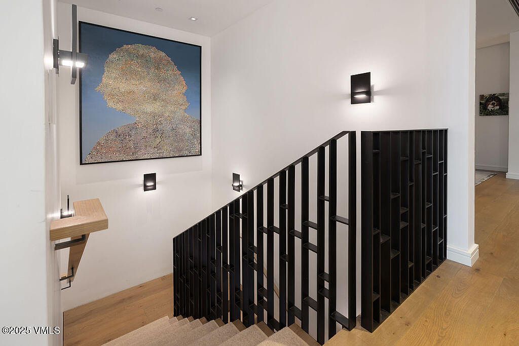 This modern staircase area features a striking black metal railing with a geometric, vertical slat design that provides a contemporary architectural focal point. The space is illuminated by minimalist wall-mounted sconces and recessed ceiling lighting, highlighting a large, textured abstract painting on the wall. Light-toned wood flooring and neutral walls create a clean, sophisticated aesthetic, while a small wooden wall-mounted shelf adds a functional, minimalist touch.