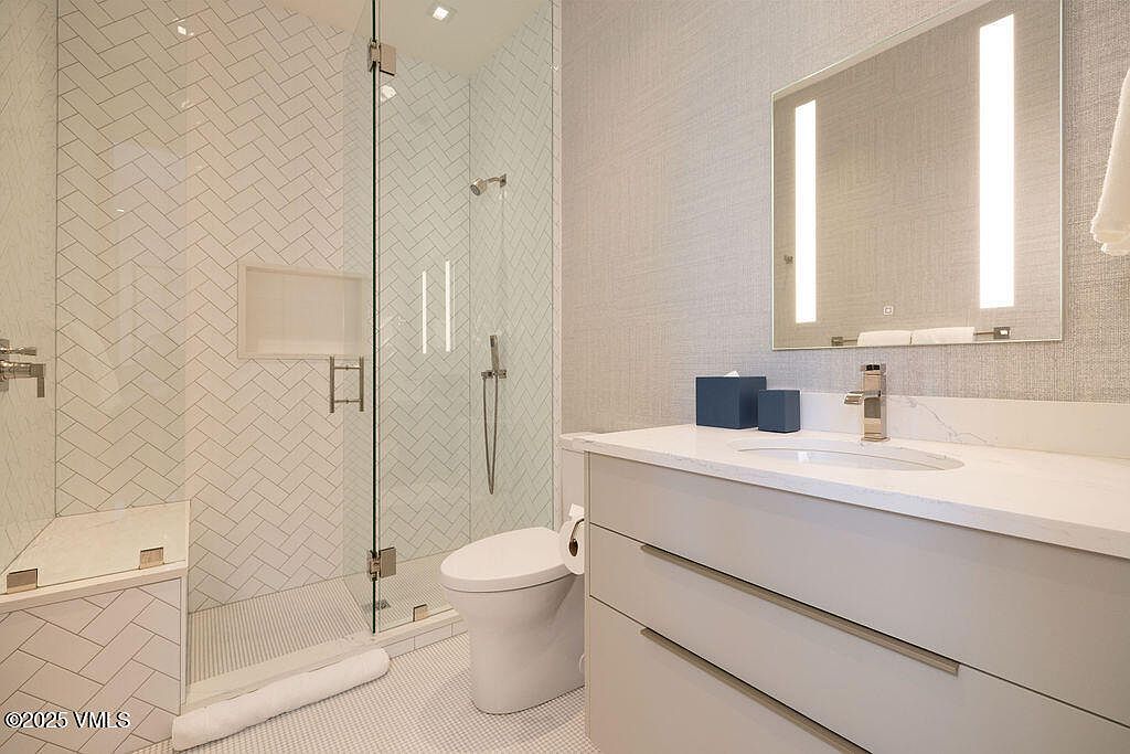 This modern bathroom features a sleek vanity with a light-toned countertop and a rectangular mirror with integrated vertical lighting. To the left, a glass-enclosed shower showcases white herringbone-patterned wall tiles and a built-in niche. The space is finished with a neutral color palette, creating a clean, contemporary, and bright atmosphere.