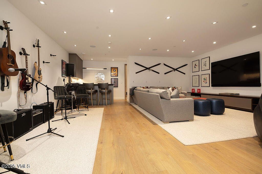 This open-concept living area features a spacious layout that seamlessly integrates a music-themed lounge space with a comfortable seating area. The room is anchored by light wood flooring and recessed lighting, with a wall dedicated to musical instruments and audio equipment on one side and a large television mounted above a low-profile media console on the other. A neutral-toned sectional sofa and dark blue ottomans create a cozy gathering spot, while a bar area with stools provides additional seating near the kitchen.