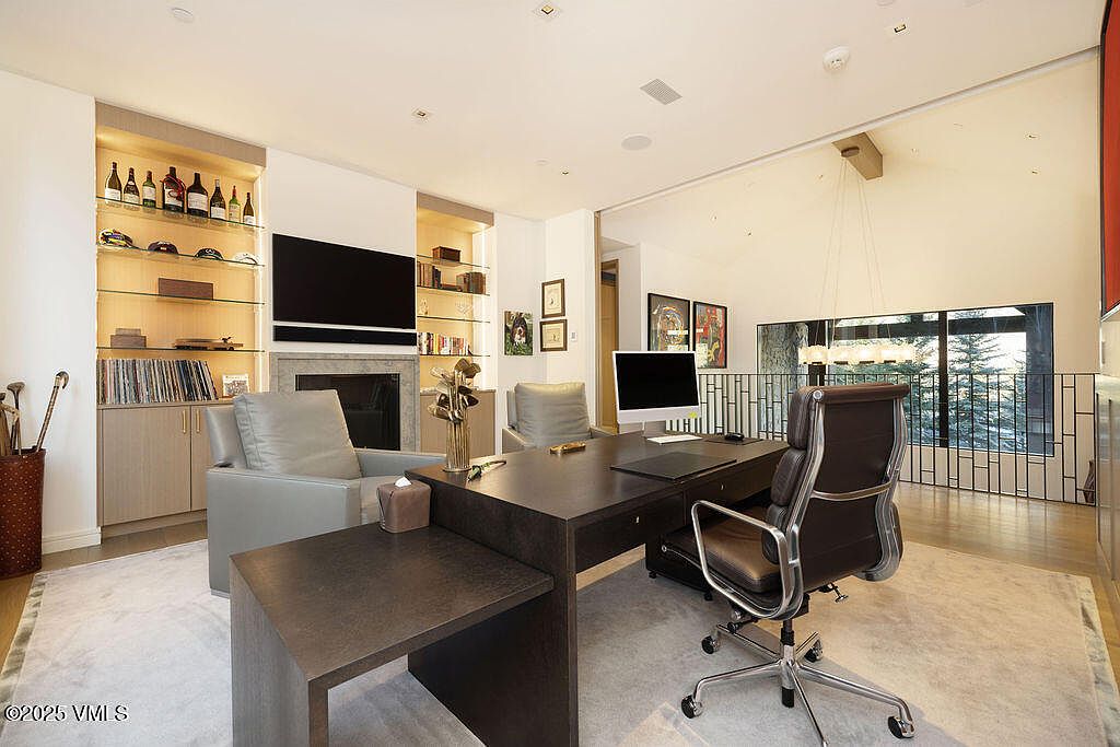 This sophisticated home office features a large, dark-toned desk positioned centrally on a light-colored area rug, complemented by a leather executive chair and two additional guest chairs. The room is anchored by a fireplace with a stone surround and built-in shelving units on either side, which are illuminated by integrated lighting. Large windows with a view of trees and a metal railing provide natural light, creating an open and professional atmosphere.