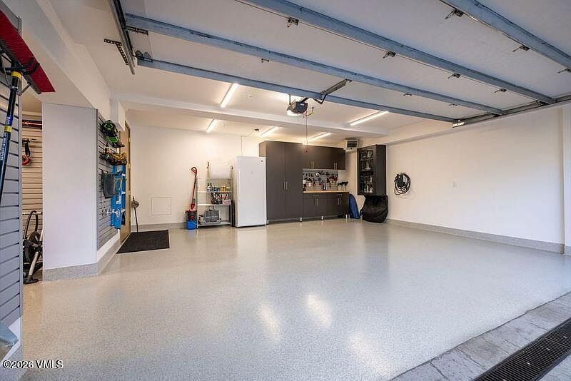 This spacious, well-organized garage features a clean, light-colored epoxy floor and bright overhead lighting. The space includes a dedicated storage area with dark cabinetry, a white refrigerator, and a metal shelving unit, alongside wall-mounted storage racks for tools and equipment. The perspective is from the garage entrance, showcasing a tidy, functional area with ample room for vehicles and organization.