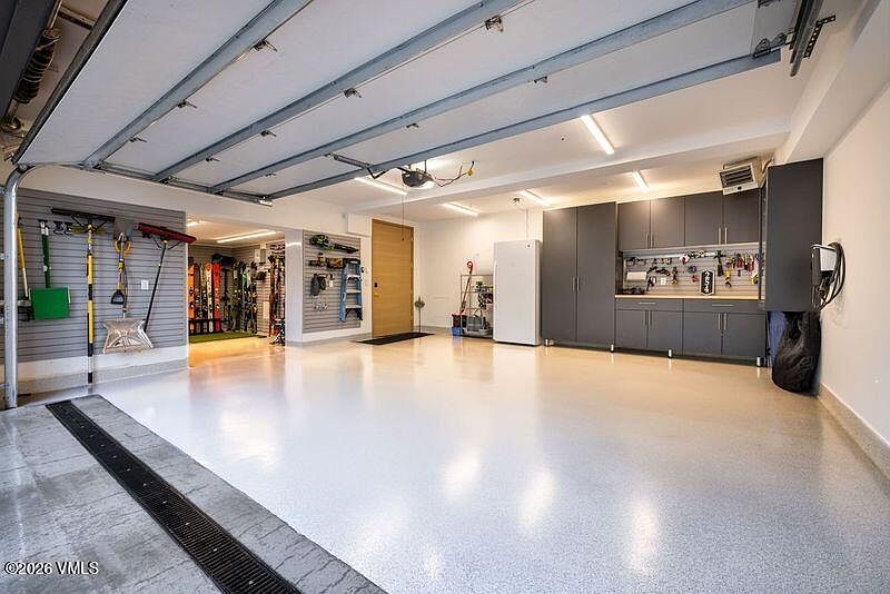 This spacious, well-organized garage features a clean, light-colored epoxy floor and a high ceiling with integrated lighting. The space is equipped with extensive wall-mounted storage systems, including cabinets, shelving, and specialized racks for skis and tools, creating a highly functional and tidy environment. The perspective is wide-angle, capturing the entire depth of the garage from the entrance, highlighting its ample storage capacity and pristine condition.