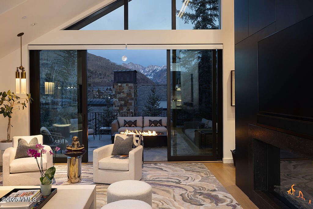 This sophisticated living room features a contemporary design with a large, patterned area rug and two plush white armchairs facing a central fireplace. Large glass sliding doors open onto a balcony, framing a stunning mountain view and a cozy outdoor fire pit area. The room is accented by a tall, dark-toned fireplace surround and elegant pendant lighting, creating a warm and inviting atmosphere.