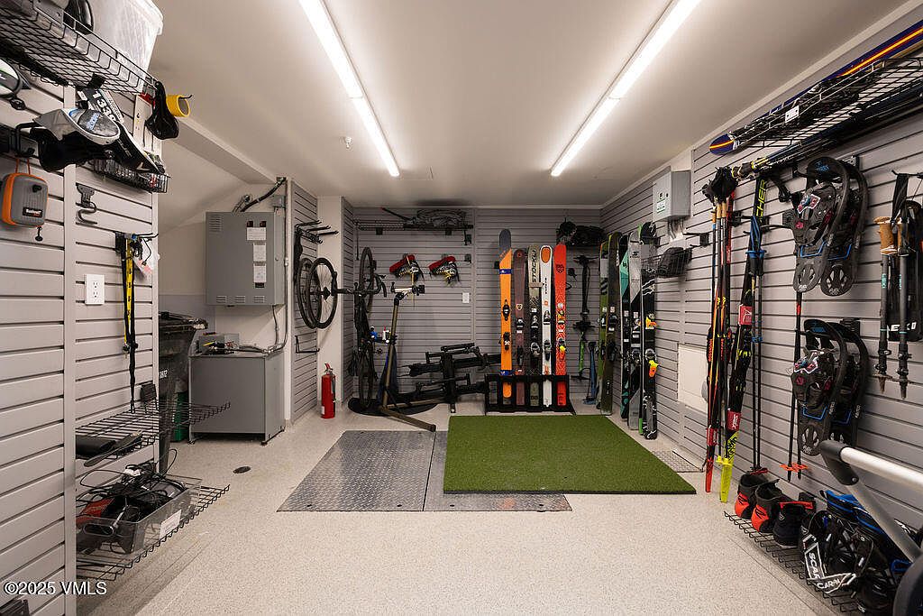 This organized garage space is designed for active storage, featuring extensive wall-mounted slat systems for hanging skis, boots, and bicycles. The room includes a central green turf mat, a metal diamond-plate floor section, and bright overhead lighting that illuminates the various sporting equipment. The space is highly functional and tailored for outdoor enthusiasts, providing a clean and efficient storage solution.