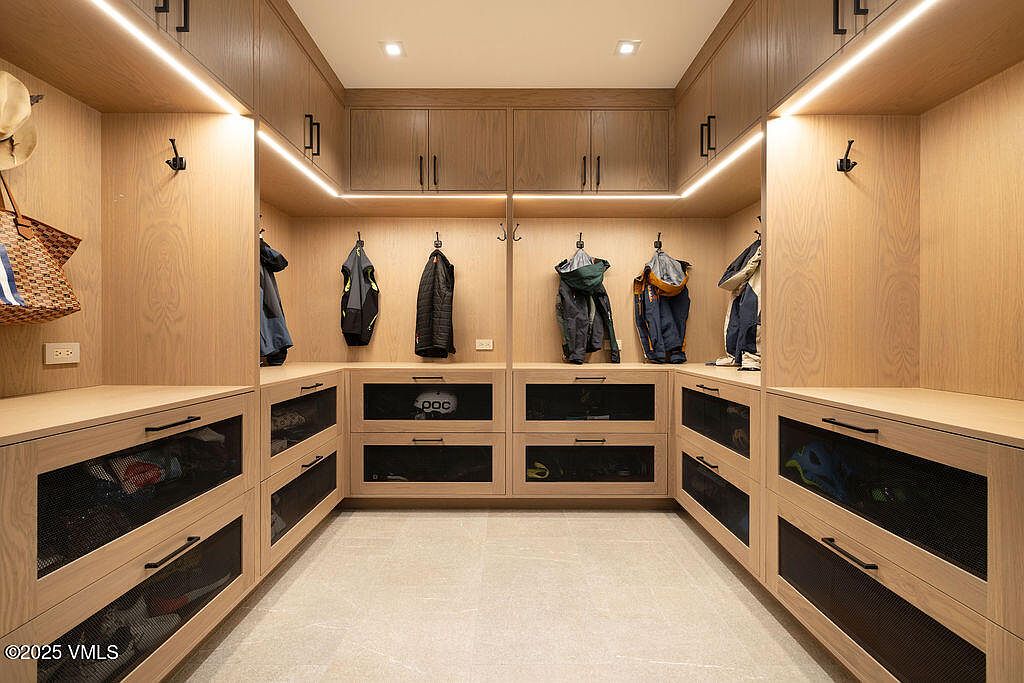 This organized mudroom or walk-in closet features custom light wood cabinetry with integrated LED strip lighting and a symmetrical layout. The space includes multiple hanging hooks for gear, upper storage cabinets, and lower drawers with mesh fronts for ventilation. The neutral stone-look tile flooring and clean lines create a functional and modern storage environment.