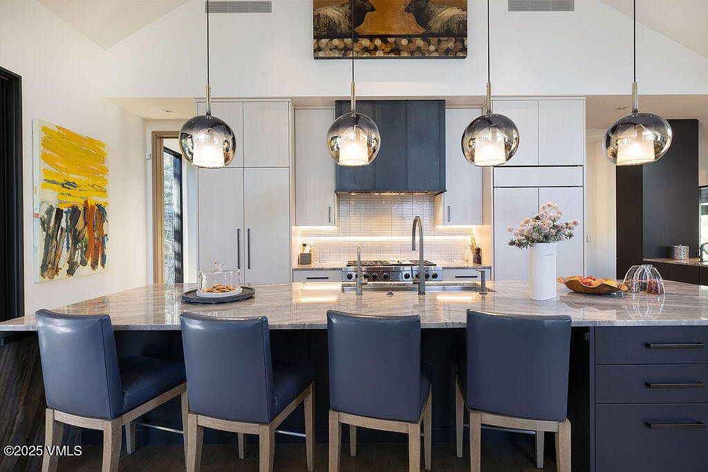 This modern kitchen features a large central island with a stone countertop and four blue upholstered chairs, serving as a primary focal point. The space is equipped with light-toned cabinetry, a dark range hood, and four spherical pendant lights hanging above the island. A vibrant abstract painting adds a pop of color to the left, while the overall aesthetic is clean, contemporary, and well-lit.