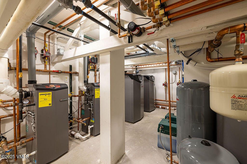 This utility room features a complex mechanical system with multiple Lochinvar boilers and large water storage tanks. The space is characterized by exposed copper piping, industrial-grade plumbing, and concrete flooring, reflecting a functional and well-maintained mechanical area. The perspective is a wide-angle shot from the center of the room, showcasing the depth and density of the heating infrastructure.