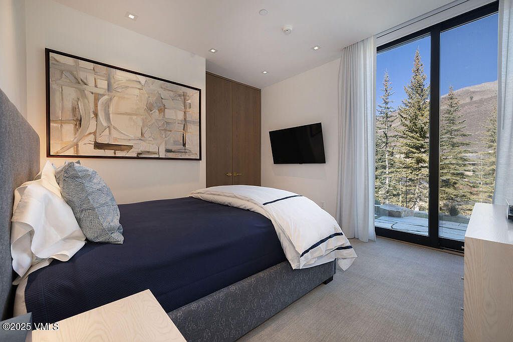 This modern bedroom features a plush bed with a dark blue coverlet and crisp white linens, positioned against a neutral wall adorned with a large abstract art piece. To the right, floor-to-ceiling glass doors provide a clear view of a serene, snow-dusted forest landscape. The room is completed by a wall-mounted television and light wood-toned closet doors, creating a clean and tranquil atmosphere.