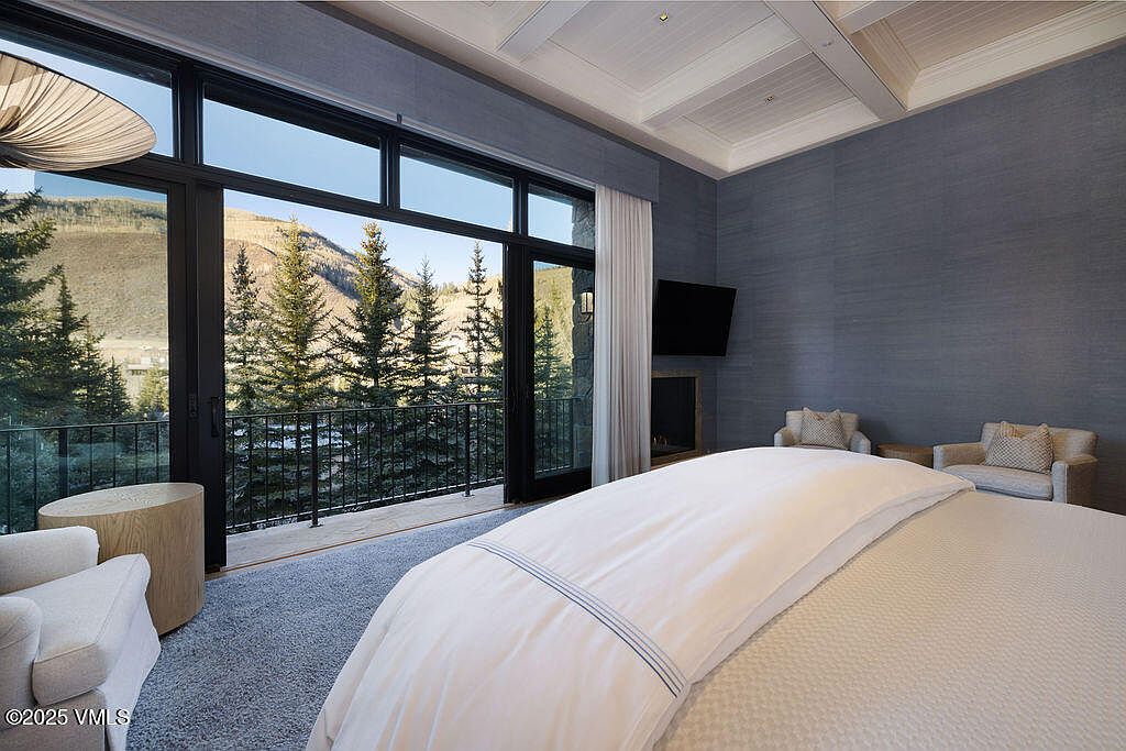 This primary bedroom features a sophisticated design with textured blue walls and a large, plush bed as the focal point. A wall of glass doors opens onto a private balcony, providing expansive views of the surrounding pine trees and mountain landscape. The room is accented by two comfortable armchairs and a minimalist fireplace, creating a serene and luxurious atmosphere.