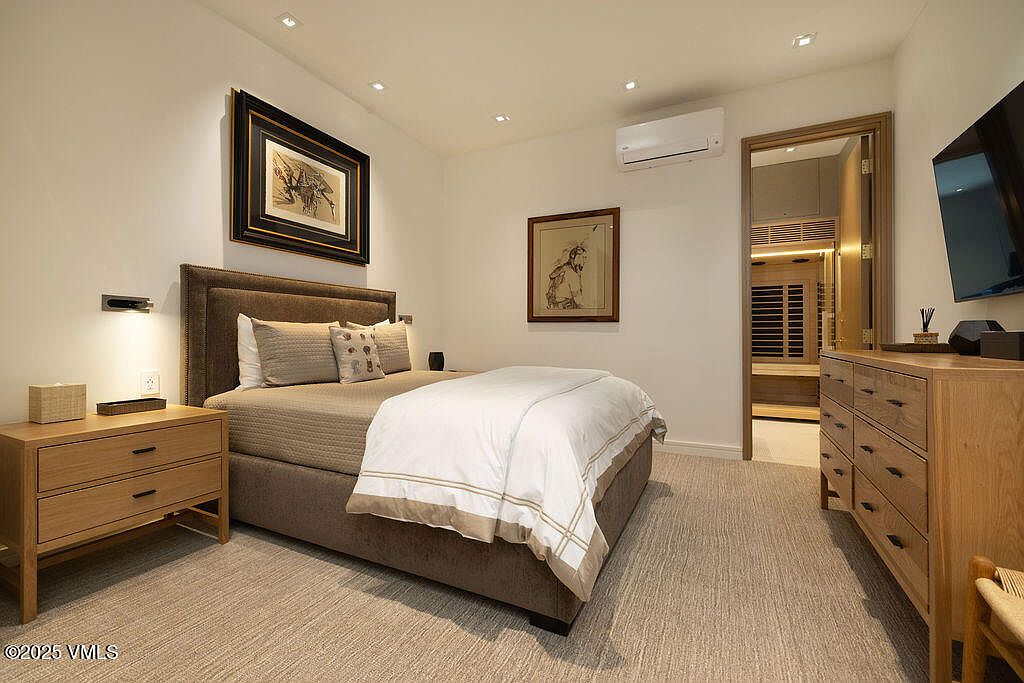 This well-appointed bedroom features a neutral color palette with a plush upholstered headboard and light wood furniture, including a matching nightstand and dresser. The room is illuminated by recessed ceiling lights and includes a wall-mounted television and a doorway leading to an adjacent sauna room. The perspective is a straight-on shot from the foot of the bed, showcasing a clean, contemporary aesthetic.