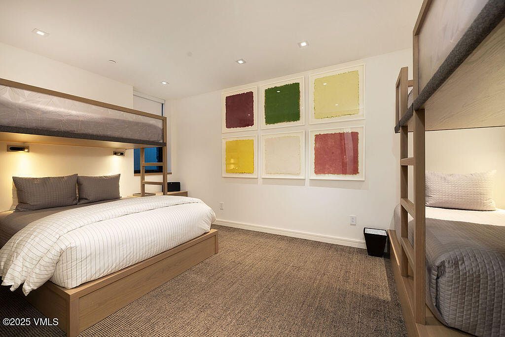 This guest bedroom features a functional layout with a bunk bed setup, including a lower queen-sized bed and an upper twin bunk. The room is decorated with a grid of six colorful, textured art pieces on the wall and is finished with neutral carpeting and light wood bed frames. The perspective is a wide-angle shot from the corner of the room, highlighting the efficient use of space for multiple occupants.