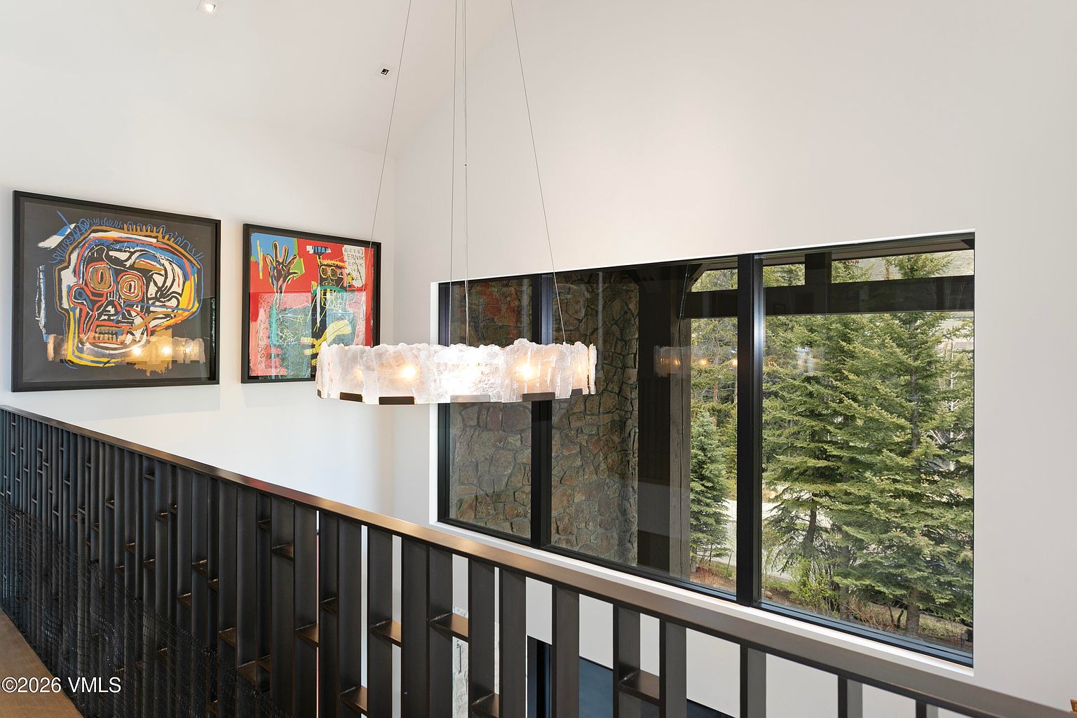 This interior view captures a landing area featuring a striking, textured circular chandelier suspended above a dark metal railing. Two vibrant, abstract art pieces hang on the white wall, while a large window provides a clear view of the surrounding pine trees and stone exterior detailing. The space feels modern and airy, with a clean aesthetic that highlights the architectural connection between the indoor and outdoor environments.