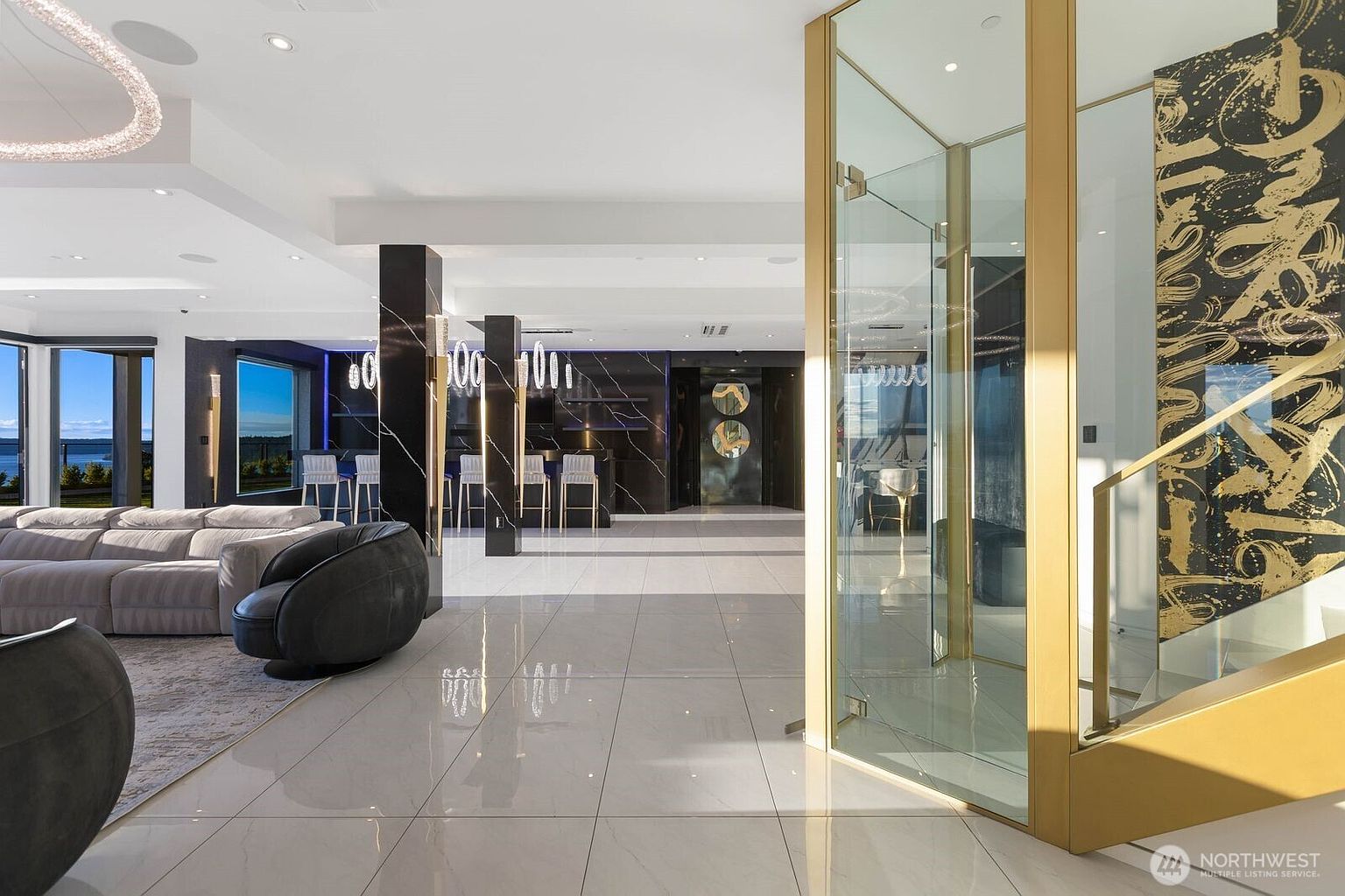 This expansive, modern living area features a large, light-colored sectional sofa and a dark, rounded accent chair set against a backdrop of high-contrast black and white decor. A prominent glass-enclosed structure with gold-toned framing stands to the right, while a sleek bar area with dark stone columns and bar stools is visible in the background. The space is illuminated by recessed lighting and a unique, sculptural light fixture, creating a sophisticated and luxurious atmosphere.