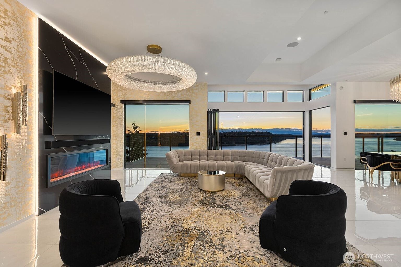 This luxurious living room features a large, curved sectional sofa and two plush black armchairs arranged around a central metallic coffee table on a textured area rug. A striking circular crystal chandelier hangs above the seating area, while a sleek fireplace is integrated into a dark, full-height stone surround. The room is defined by expansive glass walls that provide panoramic views of the water and sunset, creating a sophisticated and airy atmosphere.