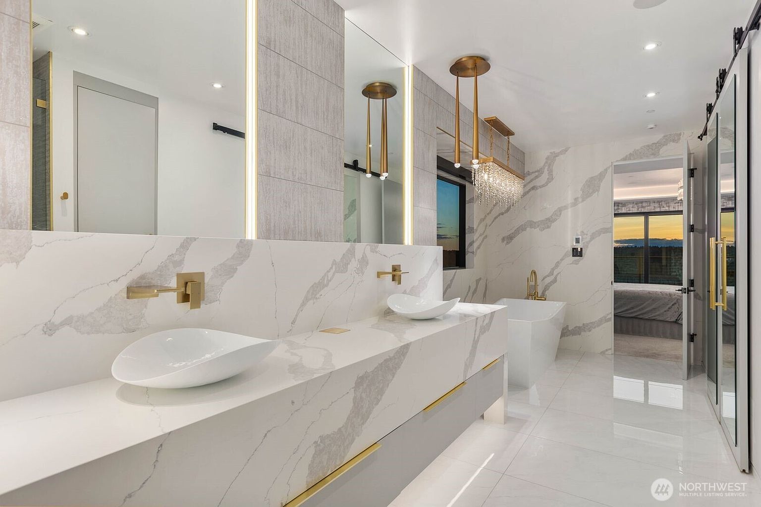 This luxurious primary bathroom features a double vanity with a long, patterned stone countertop and two vessel sinks. The space is accented with gold-toned fixtures, vertical integrated lighting, and a crystal chandelier, creating a sophisticated and modern aesthetic. A freestanding soaking tub is positioned near a large window, while a glass-enclosed shower area is visible to the right, all set against a backdrop of polished light-colored tile flooring.
