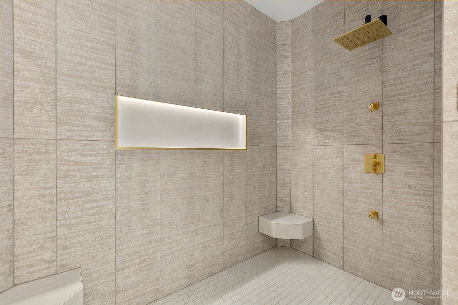 This modern walk-in shower features large-format, textured neutral tiles that create a sophisticated, contemporary aesthetic. The space is equipped with gold-toned fixtures, including a rainfall showerhead and a built-in corner bench, complemented by a recessed, illuminated wall niche. The perspective is a wide-angle view capturing the clean lines and high-end finishes of this luxurious shower enclosure.