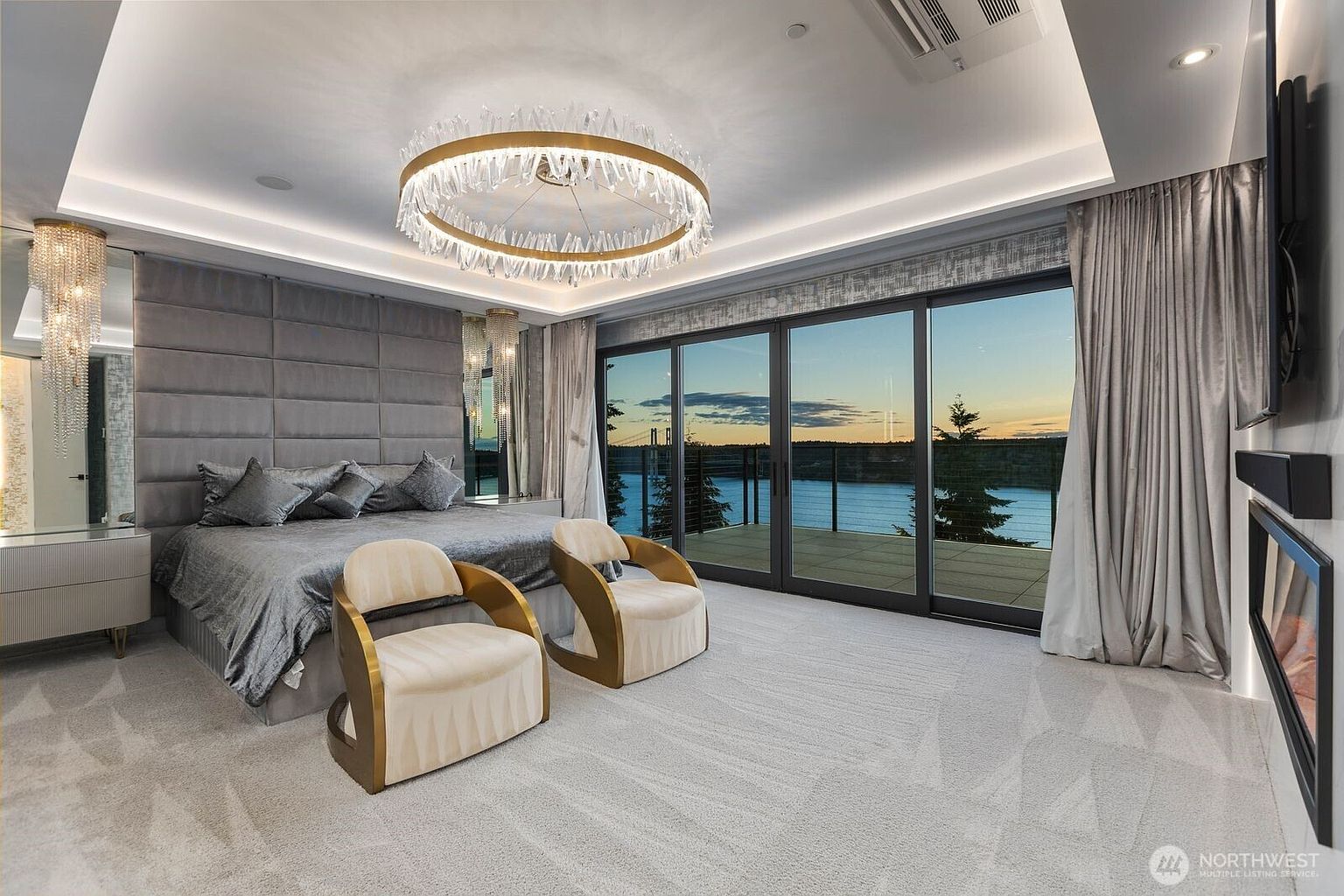 This luxurious primary bedroom features a plush, upholstered headboard wall and a sophisticated circular crystal chandelier that serves as a focal point. Two elegant, curved-back chairs with gold-toned frames sit at the foot of the bed, while large glass sliding doors provide an expansive view of the water and bridge at sunset. The room is finished with neutral carpeting and soft, floor-length drapery, creating a serene and high-end atmosphere.