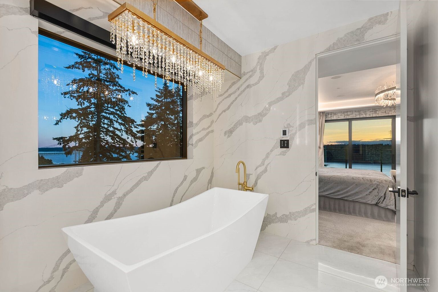 This luxurious primary bathroom features a sleek, freestanding white soaking tub positioned beneath a large window that offers a scenic view of trees and water. The walls are adorned with large-format stone slabs featuring dramatic veining, complemented by a gold-toned faucet and a sophisticated crystal chandelier. A doorway leads into an adjacent bedroom, creating a seamless flow between the private living spaces.