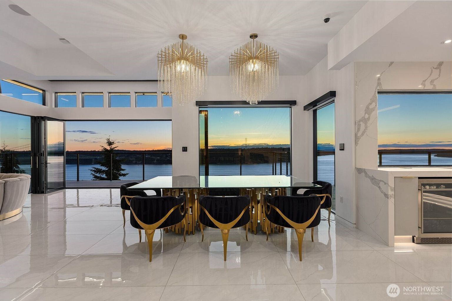 This sophisticated dining area features a glass-topped table surrounded by six modern chairs with distinctive gold-toned metal bases and black upholstery. Two ornate, tiered crystal chandeliers hang above the table, reflecting the light against the polished, light-colored tile flooring. The room is framed by expansive glass walls that provide an unobstructed, panoramic view of the water and horizon at sunset, creating a seamless connection between the interior and the outdoor landscape.