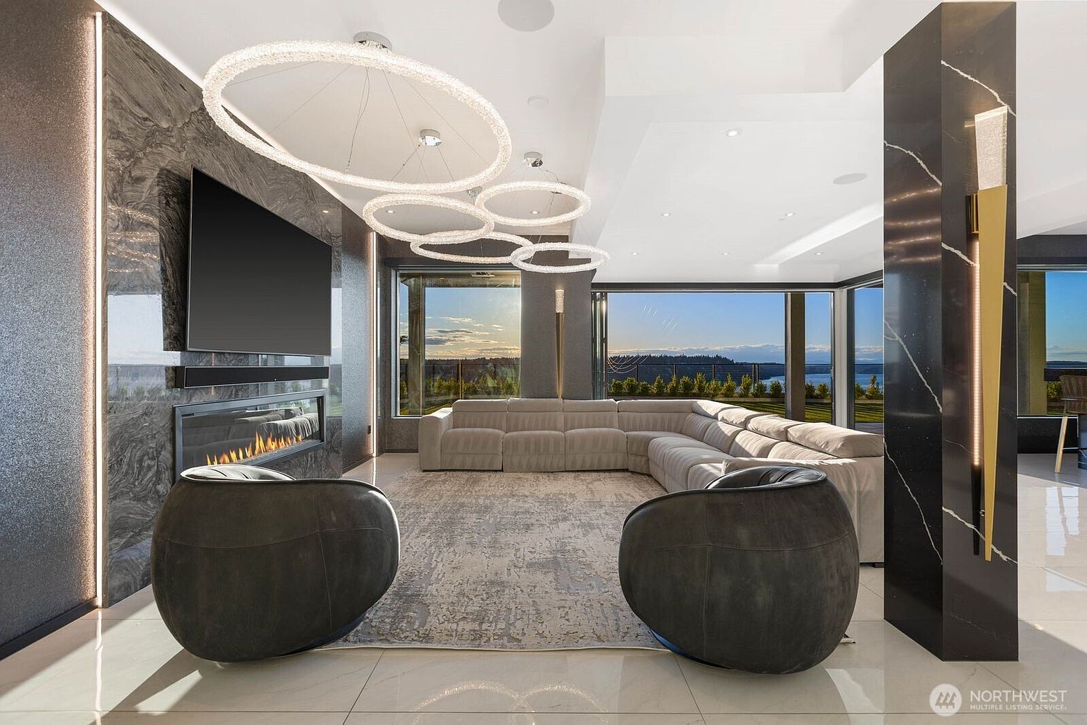 This contemporary living room features a large, light-colored sectional sofa arranged around a textured area rug, complemented by two dark, rounded accent chairs in the foreground. A sleek, linear fireplace is set into a dark, stone-clad wall that also houses a large wall-mounted television. The space is illuminated by a series of circular, modern pendant lights and benefits from expansive glass walls that provide an unobstructed view of the outdoor landscape.